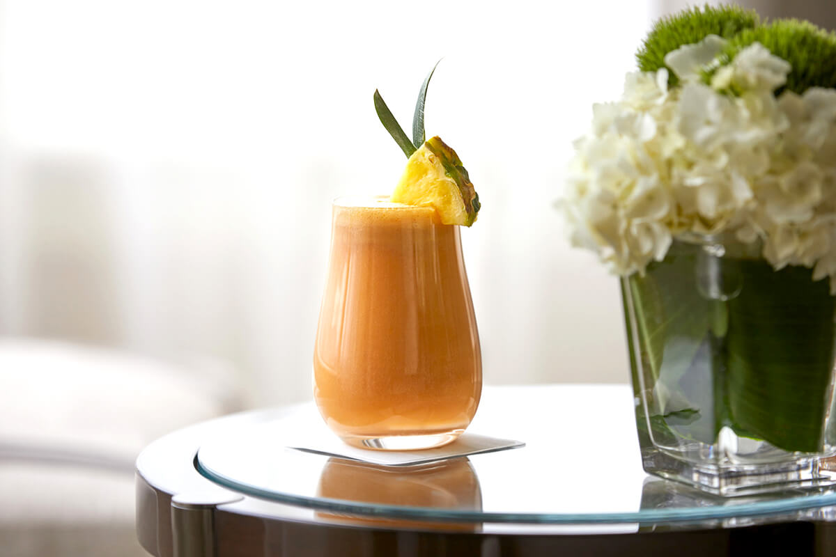 A glass filled with a colorful smoothie, placed on a table with a green leaf and a pineapple. The background shows a blurred view of the surrounding environment, which appears to be a luxurious resort or spa.